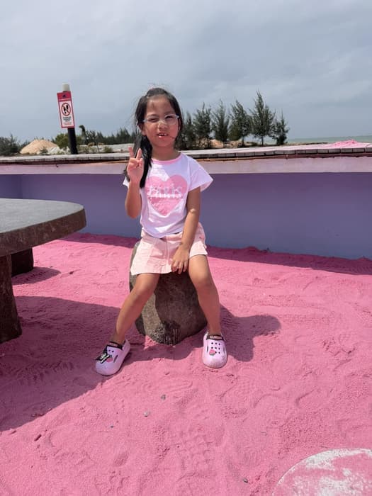 A young girl making a peace sign, sitting on the pink sand at a beach club.