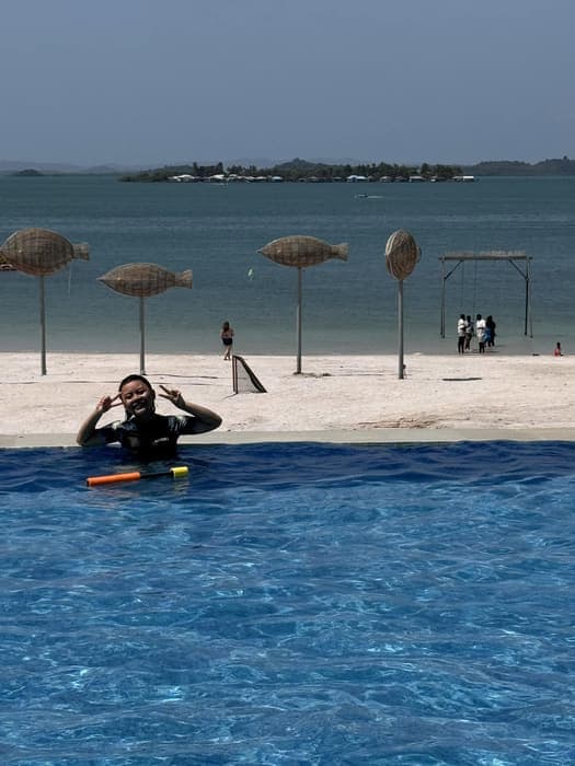 A child in the infinity pool at a beachfront resort, with woven fish sculptures on the sand behind.