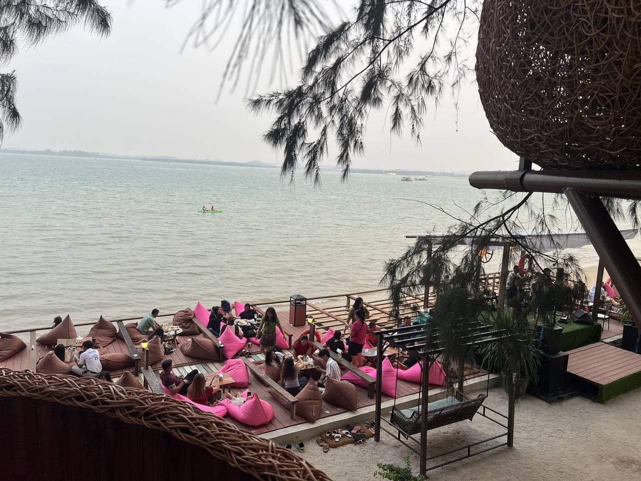 Guests on pink beanbags on a beachfront deck below a woven nest seat, the sea behind them