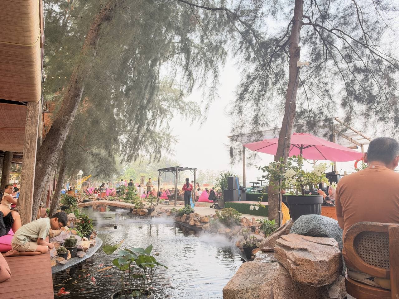 A boy watching the koi in a misty garden pond at a beachfront treehouse cafe, a pink parasol beyond